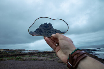 A hand holds a cloud-shaped mirror reflecting a mountain peak, with a cloudy ocean backdrop 