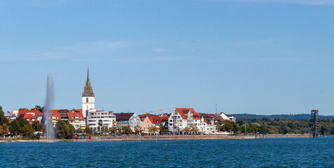 A scenic view of a lakeside fountain and a church tower in Friedrichshafen, Germany, under a clear blue sky on a sunny summer day