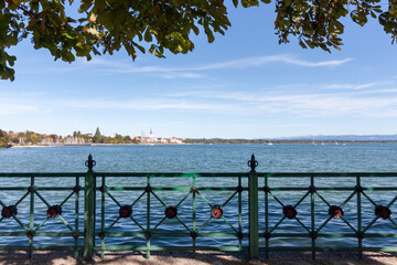 A picturesque view of a lakeside town on a sunny day, with a traditional green metal fence in the foreground and a blue lake stretching to the horizon