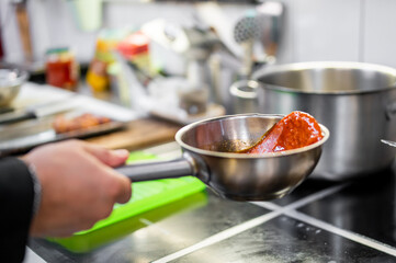 Close-up of a ladle with red sauce held over a stovetop in a professional kitchen. Cooking process with stainless steel pot, utensils, and ingredients in background