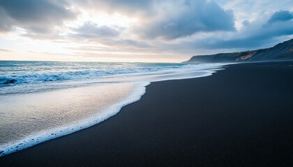 The image shows foamy ocean waves washing ashore on a dark sand beach with a rocky cliff in the background under a partly cloudy sky.