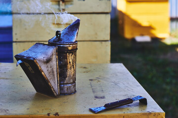Metal beekeeping smoker emitting smoke standing on yellow surface near metal hive tool with blurred...