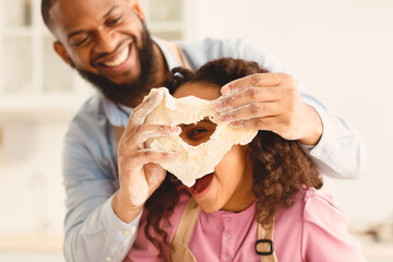 Portrait of funny black man and and cute daughter playing with food, fooling around with dough, laughing girl looking through hole, having fun in the kitchen, making pie for Father's day
