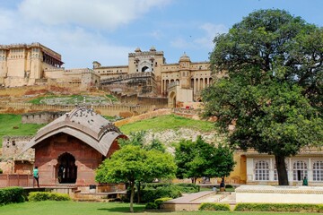 Obraz premium Amber Fort in Jaipur, India, stands prominently in the background with its grand architecture and ochre sandstone facade. In the foreground, a lush park features large, leafy trees that provide shade.
