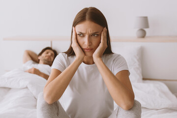 Offended sad upset pretty young caucasian woman sits on bed while husband sleeps in bedroom interior. Covid-19 pandemic, self-isolation, boredom at home, quarrel, snoring and family problems, panorama