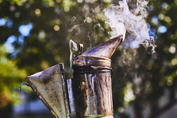 Closeup showing metal beekeeping smoker emitting smoke outdoors, device used by beekeepers for calming honey bees during hive inspection, blurred green background visible