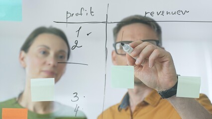 Professional team members brainstorming strategy, writing notes on transparent whiteboard with colorful sticky papers during collaborative meeting in office. Business people at work