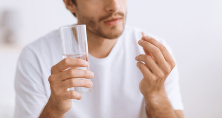 Pensive millennial european man hold glass of water and look at pill in hand, close up. Male...
