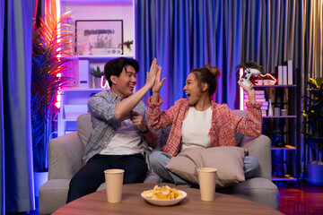 Couple gamer making high five holding joysticks be winner together complete level sitting sofa playing video game with front snack and drinks at neon light color living room at modern home. Infobahn.