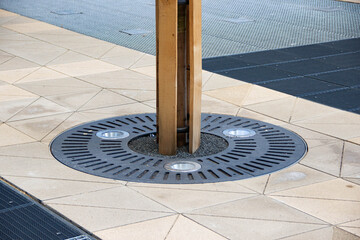 Urban tree planter with decorative metal grate and wooden support, surrounded by patterned pavement tiles, enhancing city landscape and green space design