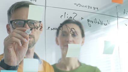 Business professionals brainstorming near glass board, using sticky notes for strategic planning, writing calculations, sharing innovative ideas during collaborative work session