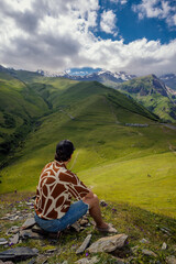 A tourist sits on a hill with his back to the green valley and looks at the Caucasus Mountains in Georgia