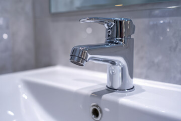 Close-up of a modern chrome faucet mounted on a white ceramic sink with blurred grey tiled wall in a contemporary bathroom setting