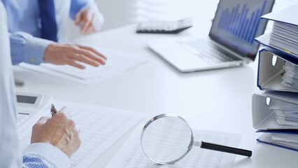 Accountants are analyzing financial reports with magnifying glass and documents, performing accounting and auditing tasks in the office, close up. Audit and taxes in business