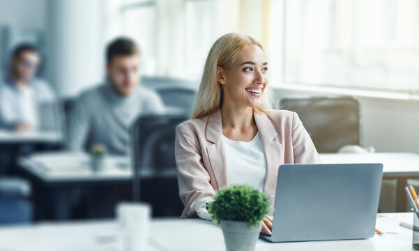 Work after lockdown. Happy young woman in suit looks out the window and works at laptop in office interior with plant in pot and antiseptics during coronavirus outbreak, free space