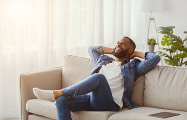 Obraz premium Happy satisfied relaxed young black bearded male resting in living room interior with tablet on sofa. Break after work in own apartment, thinking about new business ideas at home, self-isolation