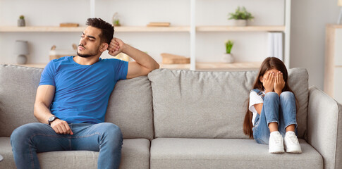 Young father sitting separate on couch with offended crying little kid girl in tears, man ignoring...