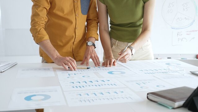 Two business professionals are examining financial data and reports, pointing at charts and graphs displayed on a table during a collaborative office meeting - Powered by Adobe