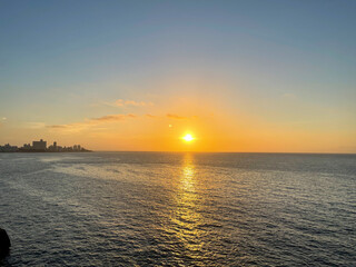 Golden sunset over Havana coastal horizon, warm sunlight reflecting across the calm ocean waters, distant city skyline