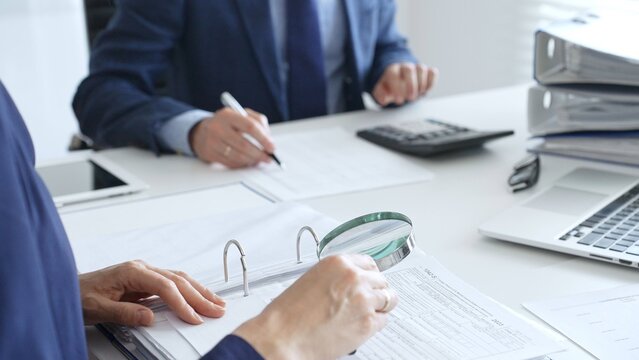 Close up of female accountant hands using calculator and magnificent glass while collaborating with male colleague in office, calculating taxes and analyzing financial data. Audit and taxes in