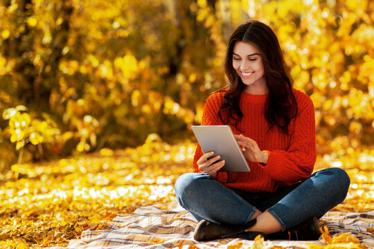 Pretty millennial woman in warm autumn outfit browsing internet on tablet computer at park. Lovely lady using touch pad to communicate online outdoors on sunny fall day