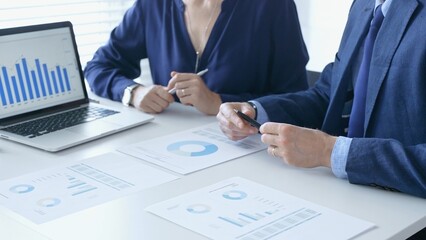 Businessman and businesswoman are analyzing financial data on a laptop and printed documents, pointing at charts and graphs during a meeting in bright office. Business people and finance concept