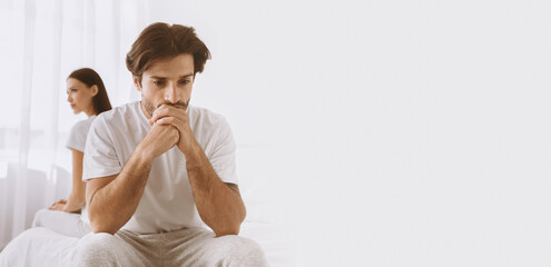 Depressed young caucasian husband with stubble sits on bed and sad, ignoring offended lady in...