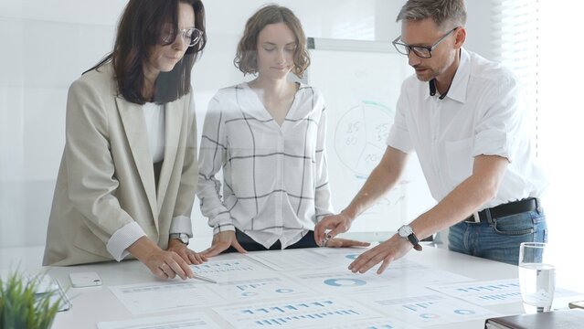 Business team collaborating on financial data analysis, pointing at charts and graphs spread out on a light table during a corporate meeting. Business people concept