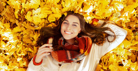 Gorgeous young woman lying among yellow autumn leaves, looking at camera and smiling, top view. Overhead shot of pretty millennial lady resting in fall foliage, enjoying change of seasons