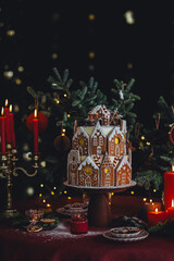 Fototapeta premium Elegant luxury Christmas table setting for family home party with homemade big gingerbread house, holiday celebration. Red tablecloth, candles, chandelier. Romantic candlelit dinner