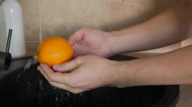Male hands diligently wash a vibrant, bright orange fruit under clear and flowing water, highlighting the significance of hygiene and food before eating or preparing any type of food for consumption