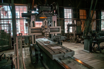 A large vintage milling machine sits prominently in an industrial workshop, surrounded by various tools and equipment. Sunlight streams through the windows, illuminating the workspace.