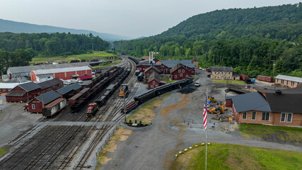 A vintage railroad station sits nestled among green mountains. Several trains are parked along the tracks. The area is quiet except for the distant sounds of nature.