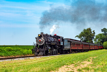 A classic steam locomotive chugs along a green valley, releasing puffing clouds of smoke into the clear blue sky.