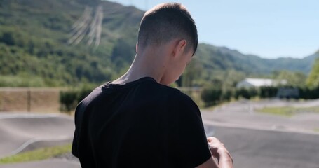 Slow-motion shot of a teen boy putting on a protective helmet at an outdoor pump track, back view.