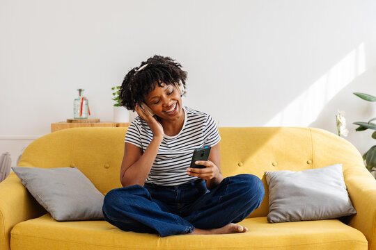 Young happy black woman sitting on a yellow couch at home, wearing headphones and enjoying music from her smart mobile phone. She is smiling and having a good time