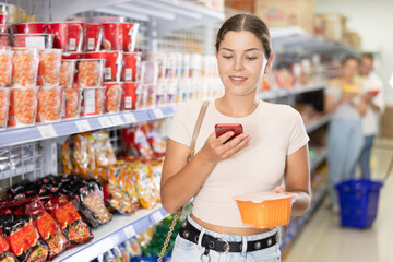 Casual interested young girl using smartphone to scan QR code on cup of instant noodles to get...