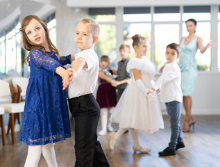 Obraz premium Smiling junior schoolchildren in party dresses learning dancing waltz with pedagogue in social hall
