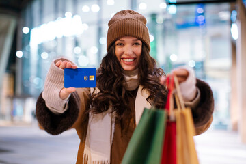 Happy woman stretching hands with credit card and shopper bags, standing outside near shopping mall. Cashback, contactless payment