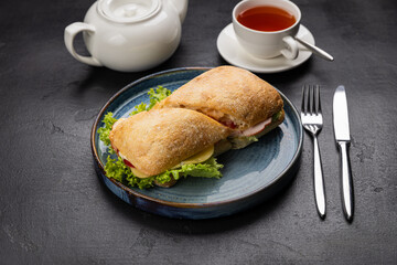 Ciabatta sandwich with cheese, ham and lettuce served with tea on a rustic black background