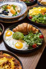 Healthy breakfast with croissant, avocado, sunny side eggs and salad on dark background