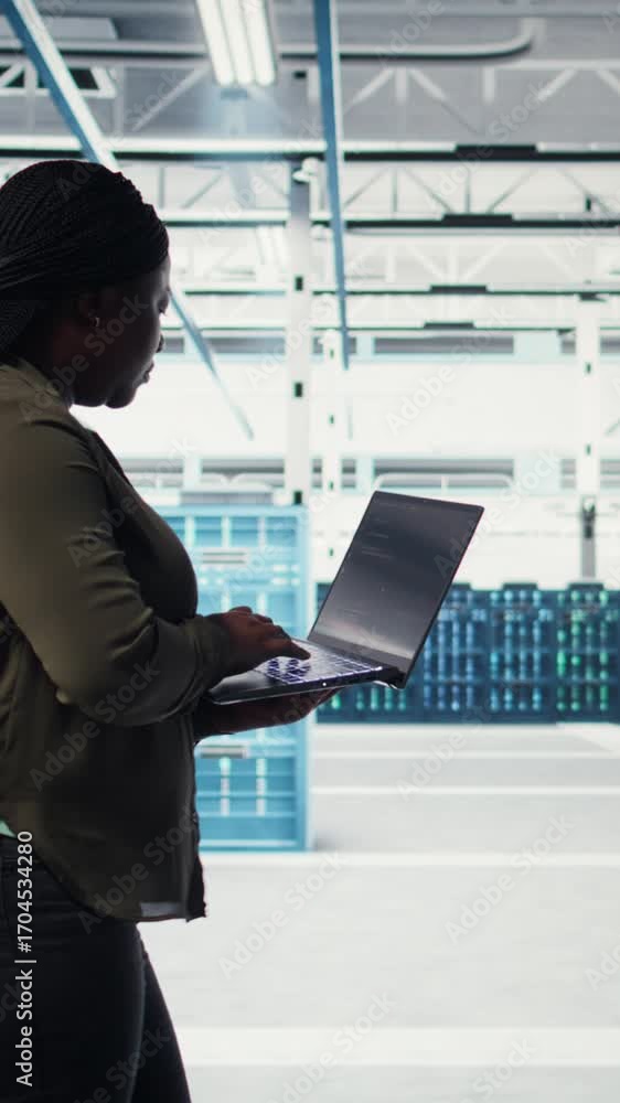Poster vertical video technician with laptop walks through data center server rows, implementing advanced p