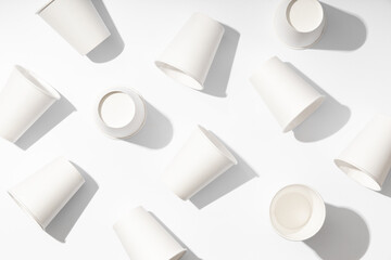 Pattern of several white paper cups for drinks on white background with hard light. Ecological disposable tableware for coffee, tea or water. Taken from a top view and flat lay.