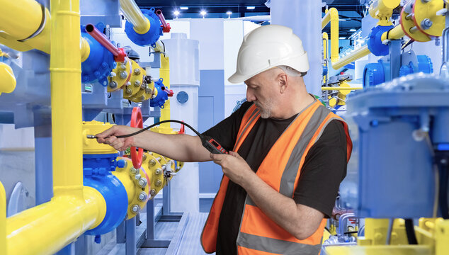 Engineer in safety vest and helmet uses gas analyzer to check pipelines and valves for leaks at industrial gas facility. Technical inspection ensures safety and system reliability.