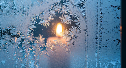 Warm candle flame glowing through frosted window pane with intricate ice crystal patterns on cold winter day creating cozy atmosphere and serene mood