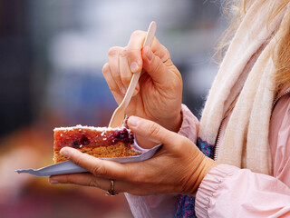 Close-up of a woman holding a paper tray with fruit dessert topped with shredded coconut, using a wooden fork to eat, at farmers market on Naplavka in Prague, Czech Republic.