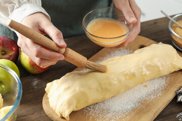 Woman spreading egg yolk onto raw apple strudel at wooden table in kitchen, closeup