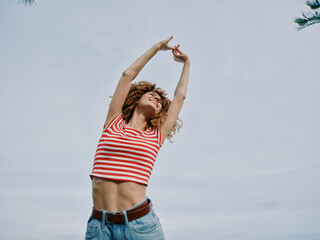 A joyful young woman stretches arms overhead under a bright sky, wearing a striped crop top and...