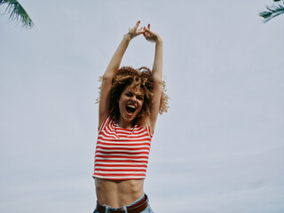 A lively young woman with curly hair raises her arms in celebration against a clear blue sky,...