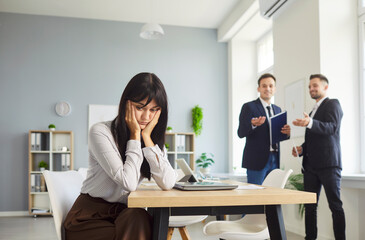 Thoughtful female employee sits alone at a desk while two coworkers chat in the background, hinting at gossip and exclusion in an office. She struggles with isolation, stress, and anxiety.